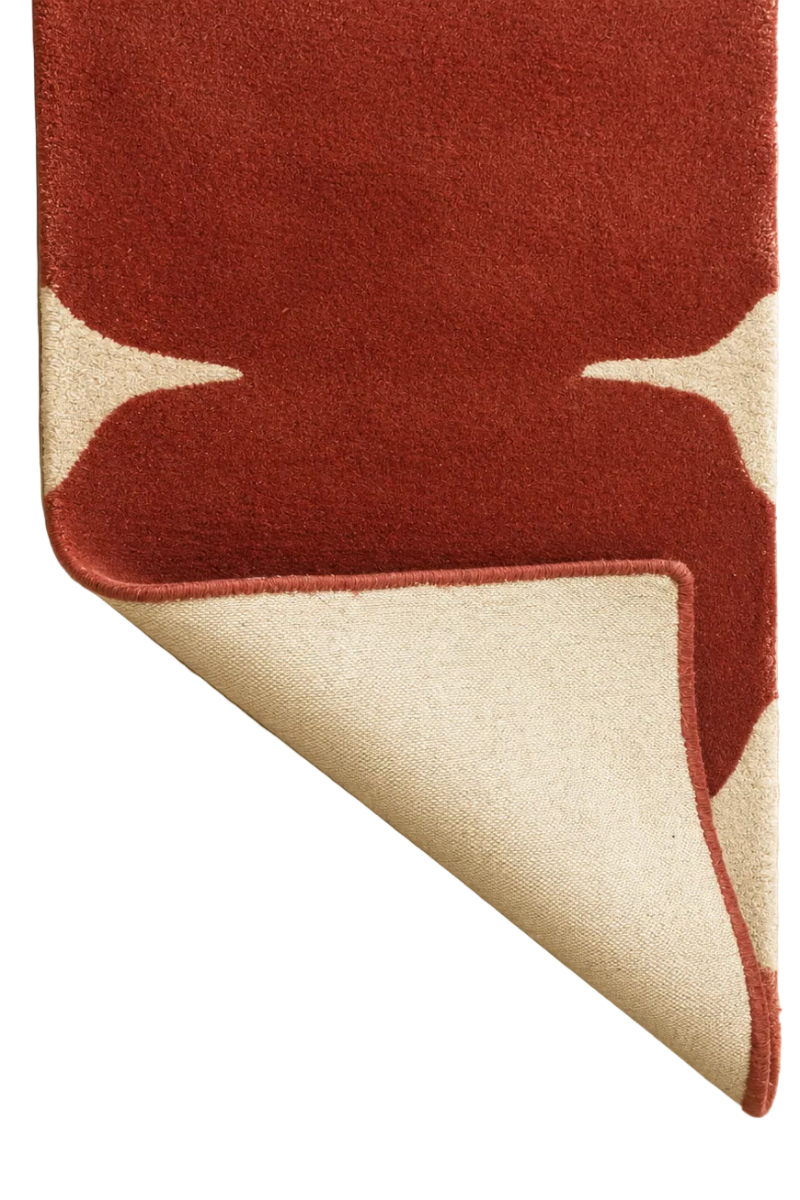A close-up of a textured area rug with an abstract organic pattern in rust red and cream, with one corner folded over to show the backing on a white background.