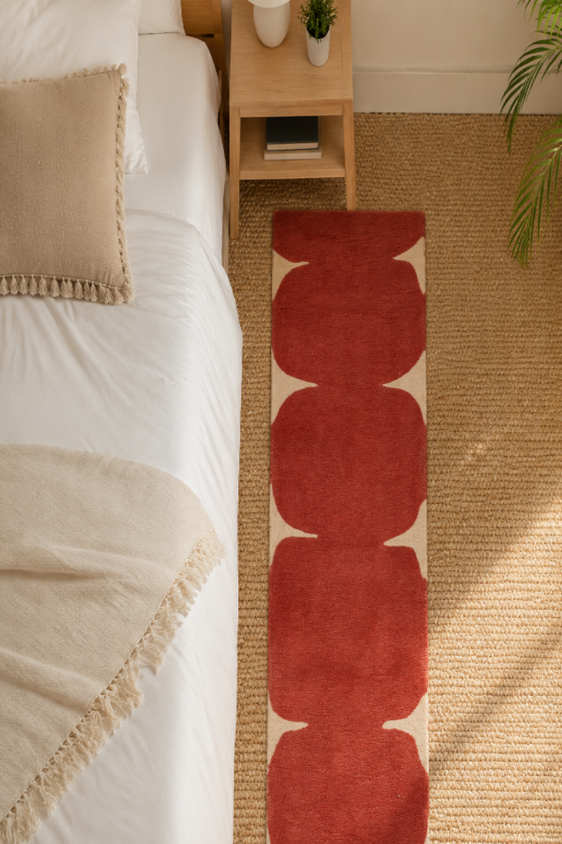 A red and cream runner rug with a repeating scallop pattern, styled on a woven jute floor between a bed and a potted plant.
