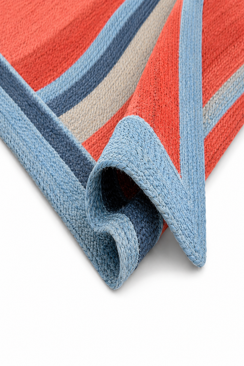 A close-up of a woven textile, likely a blanket or towel, featuring a striped pattern in coral red, light blue, navy, and beige, with a rolled corner showing the texture on a white background.