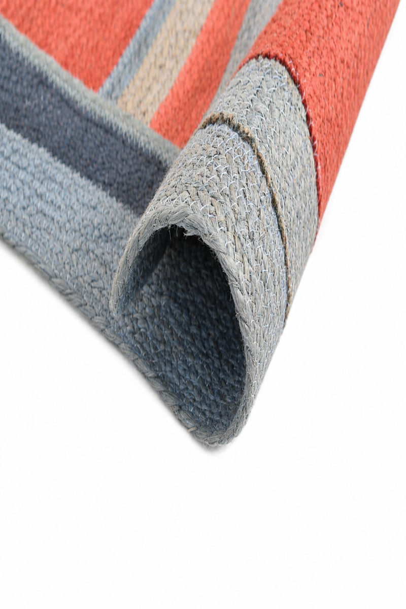 A close-up of a striped woven rug with a rolled corner, showing a textured surface in coral red, slate blue, gray, and beige stripes on a white background.
