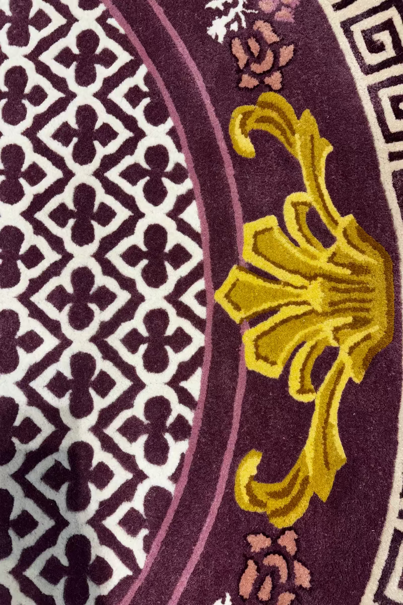 Close-up of an ornate patterned rug featuring a deep purple field with cream geometric motifs, a curved cream border, and a large raised gold floral accent.