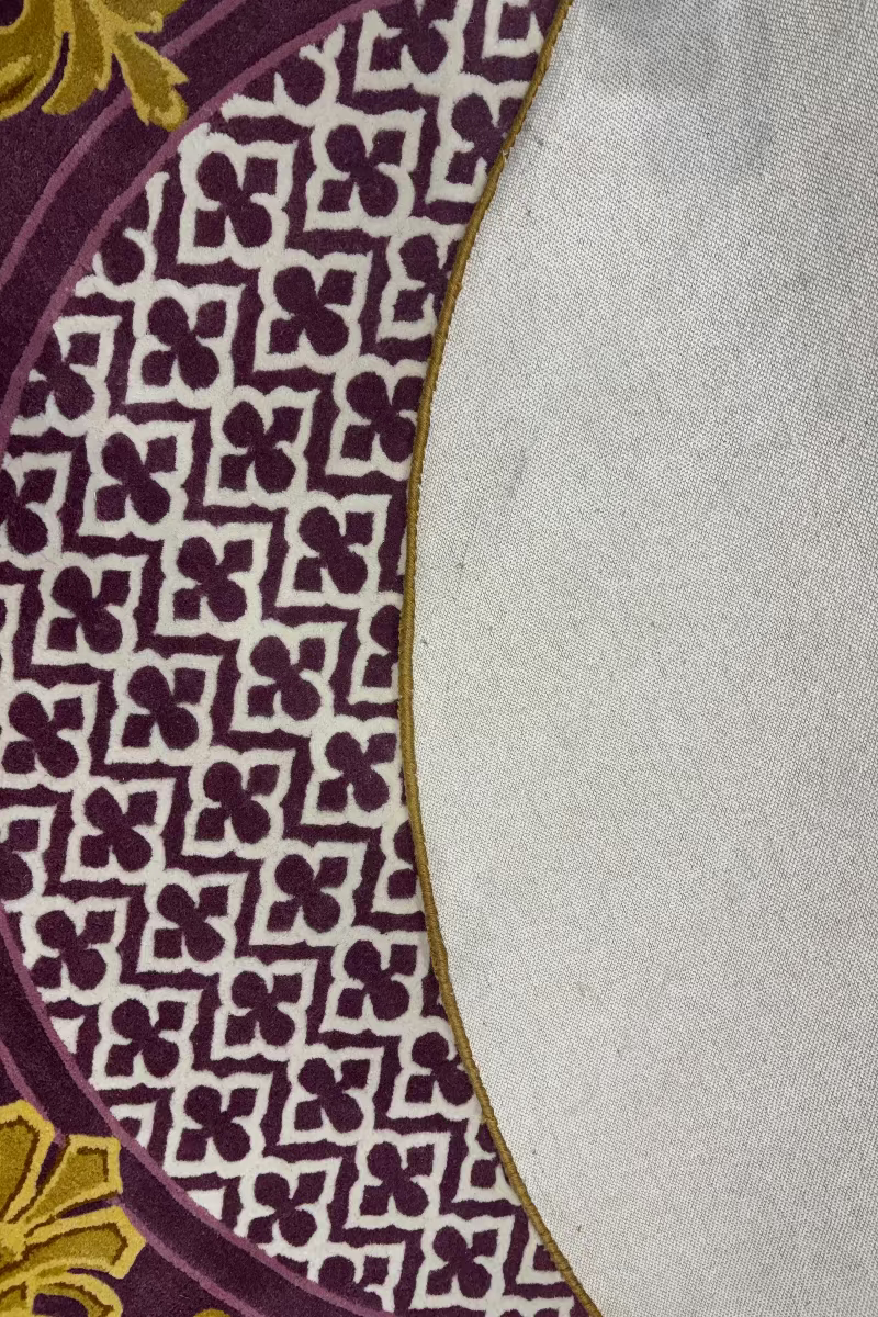Close-up of a patterned textile showing a cream background with a repeating dark purple geometric floral motif and a gold curved border, alongside a plain light gray fabric section.