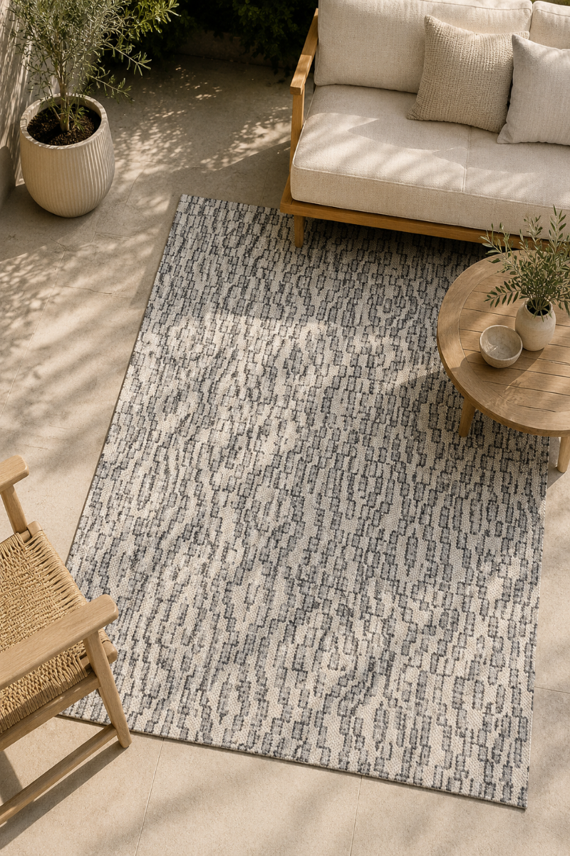 A rectangular outdoor area rug with a gray and off-white abstract brushstroke pattern, styled on a patio with a wooden sofa, chair, and round coffee table.